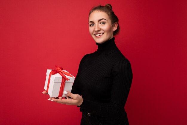 Hermosa mujer morena joven feliz aislada sobre la pared de colores de fondo vistiendo ropa casual elegante con caja de regalo y mirando a la cámara.