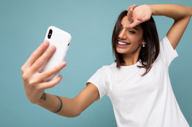 Hermosa mujer morena joven feliz sonriente con camiseta blanca casual aislada sobre fondo azul de pared sosteniendo y usando el teléfono móvil tomando selfie mirando la pantalla del gadjet.