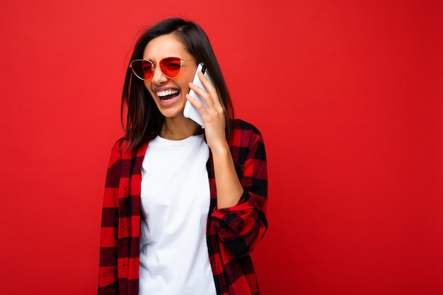 Hermosa mujer morena joven sonriente feliz positiva vistiendo elegante camisa roja camiseta blanca y gafas de sol rojas aisladas