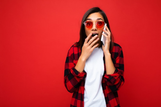 Hermosa mujer morena joven sorprendida sorprendida con elegante camisa roja camiseta blanca y gafas de sol rojas aisladas sobre pared roja hablando por teléfono móvil.