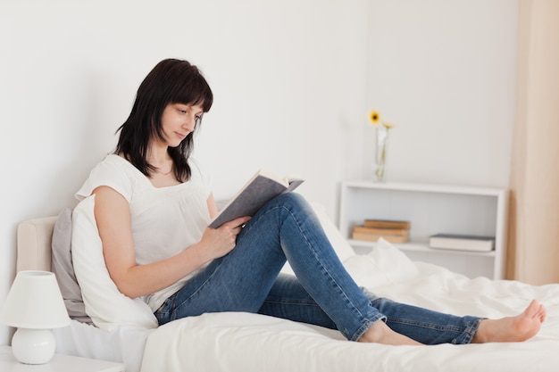 Hermosa mujer morena leyendo un libro mientras está sentado en una cama