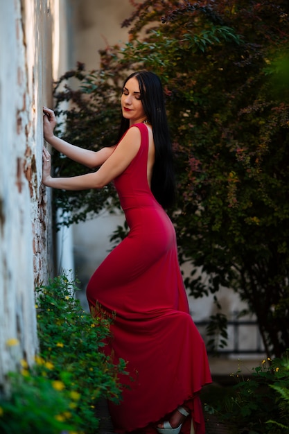 Hermosa mujer morena con pelo corto bob rizado, maquillaje delicado y labios rojos en vestido corto rojo en el parque. Poses sensuales de moda en el evento de verano. en el aire libre