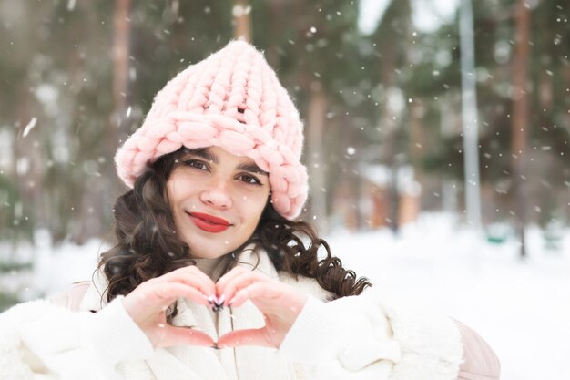 Hermosa mujer morena rizada que muestra el signo del corazón en el bosque durante las nevadas. Espacio vacio