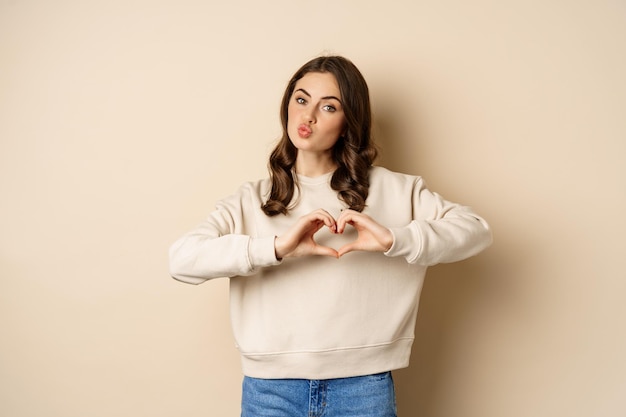 Hermosa mujer morena romántica que muestra signo de amor de corazón cuidar de ti sonriendo a la cámara de pie sobre...