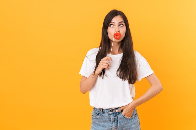 Hermosa mujer morena con ropa básica divirtiéndose y sosteniendo labios de papel aislados sobre pared amarilla