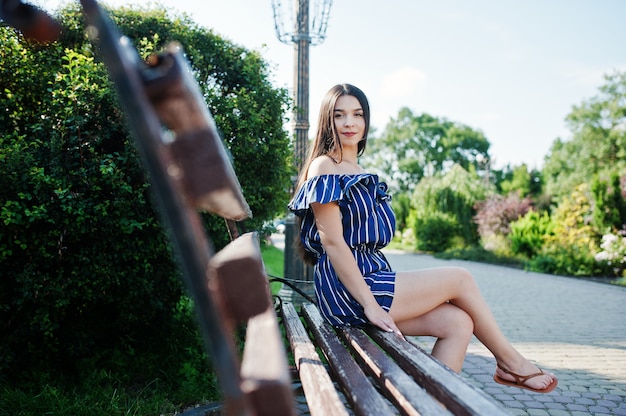 Hermosa mujer morena sentada en el banco en la calle de la ciudad usar en vestido azul a rayas.