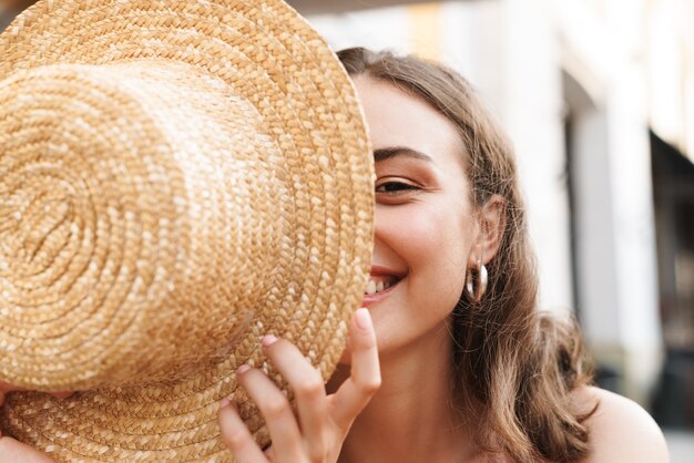 Hermosa mujer morena sonriendo y sosteniendo el sombrero de paja mientras está sentado en la calle café de verano con café para llevar