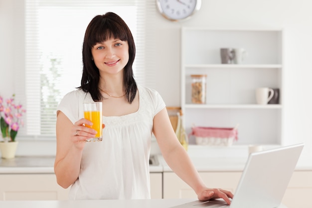 Hermosa mujer morena sosteniendo un vaso de jugo de naranja mientras se relaja con su computadora portátil