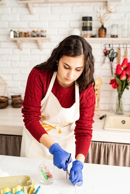 Hermosa mujer morena en suéter rojo y delantal blanco para colorear huevos de pascua en la cocina