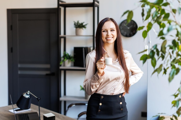 Foto hermosa mujer morena tomando un café por la mañana de pie cerca de su escritorio en la oficina mujer joven tomando café en el lugar de trabajo