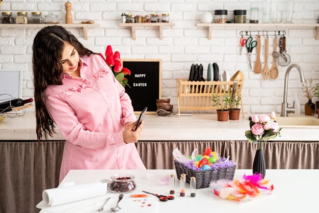 Hermosa mujer morena tomando fotos de huevos de pascua decorados mediante teléfono móvil en la cocina