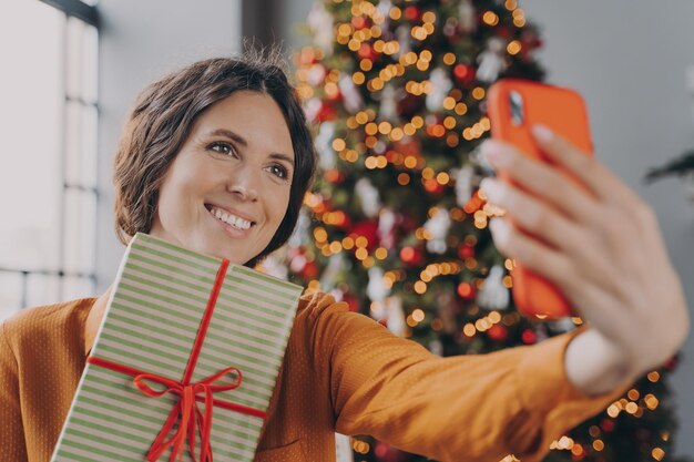 Hermosa mujer morena tomando selfie con caja de regalo de Navidad cerca del árbol de Navidad decorado en casa