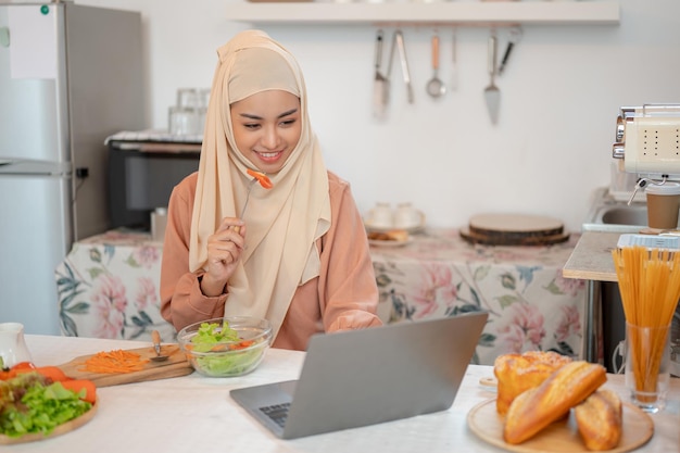 Una hermosa mujer musulmana asiática comiendo su saludable y delicioso cuenco de ensalada mientras usa su portátil