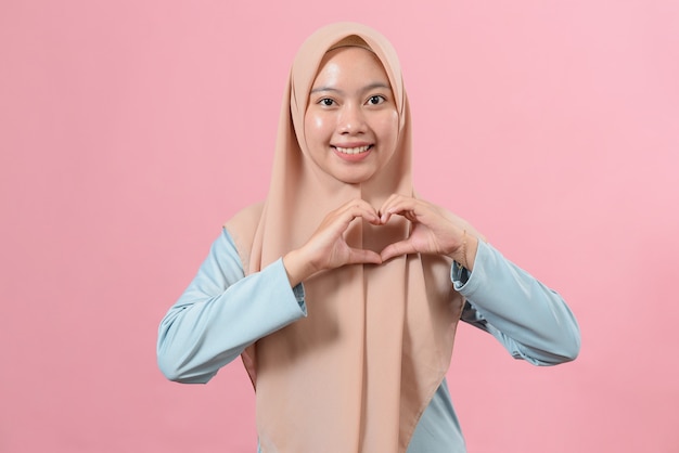 Hermosa mujer musulmana asiática hace un gesto en forma de corazón, expresa amor, dice sé mi amor, sonríe positivamente, usa hijab, posa sobre un fondo rosa. Concepto de lenguaje corporal