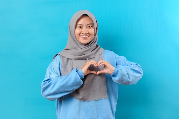 Hermosa mujer musulmana asiática hace un gesto en forma de corazón expresa amor dice ser mi amor sonríe positivamente usa poses de hiyab contra el fondo azul