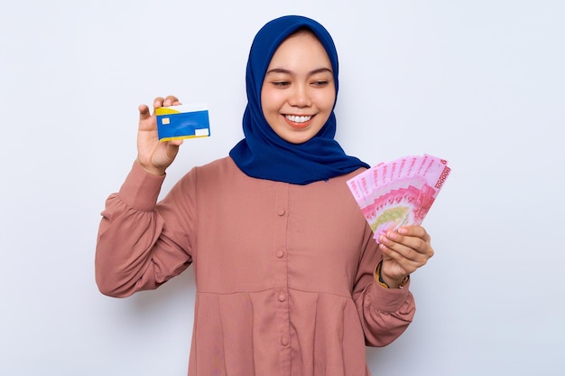 Hermosa mujer musulmana asiática sonriente con una camisa informal y un hiyab sosteniendo billetes en rupias indonesias que muestran una tarjeta de crédito aislada sobre fondo blanco Concepto de estilo de vida religioso de la gente