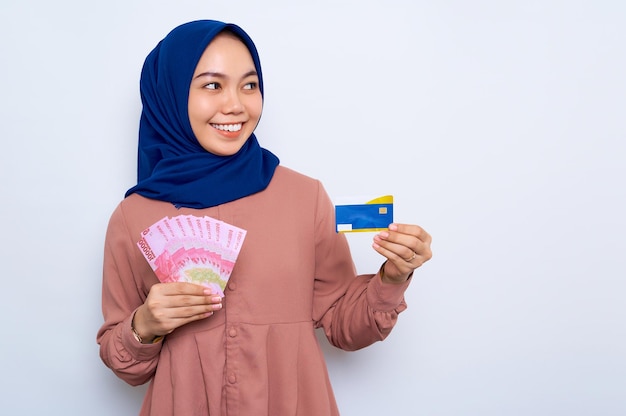 Hermosa mujer musulmana asiática sonriente con una camisa informal y un hiyab sosteniendo billetes en rupias indonesias que muestran una tarjeta de crédito aislada sobre fondo blanco Concepto de estilo de vida religioso de la gente