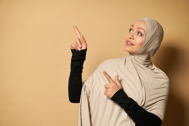 Hermosa mujer musulmana en hijab apuntando sus dedos índices hacia el lado de la superficie beige con espacio de copia