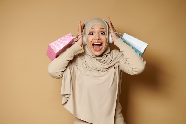 Una hermosa mujer musulmana con un hijab con bolsas de color rosa y azul en sus manos sostiene su cabeza con sus manos y grita al frente