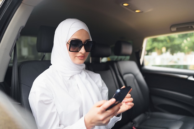 Hermosa mujer musulmana con hiyab blanco sentado en el asiento trasero de un coche y con smartphone