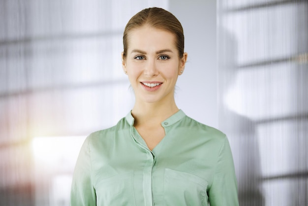 Hermosa mujer de negocios adulta vestida con una blusa verde parada directamente en una oficina soleada. Foto de negocios en la cabeza.