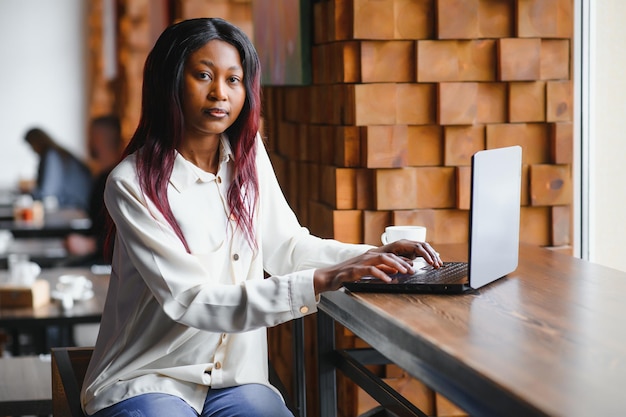 Hermosa mujer de negocios africana esperando la reunión en un espacio de trabajo conjunto y sentada con la computadora portátil. Chica hipster está navegando por la web en una computadora portátil durante el almuerzo.