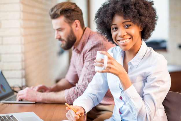 Hermosa mujer de negocios africana y hombre caucásico trabajando junto con computadoras portátiles y tazas de café cerca de la ventana en el café u oficina