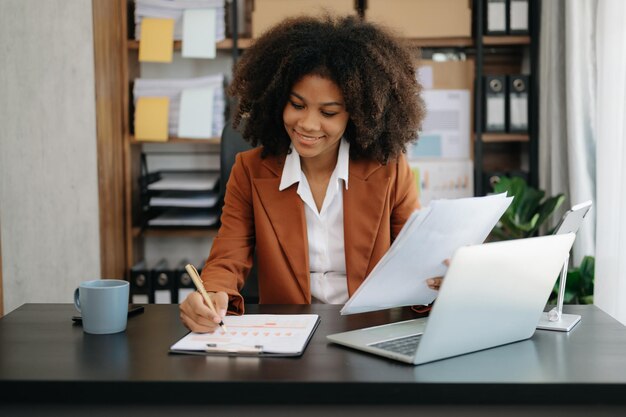 Hermosa mujer de negocios africana segura escribiendo en una computadora portátil y una tableta digital mientras sostiene café en una oficina moderna