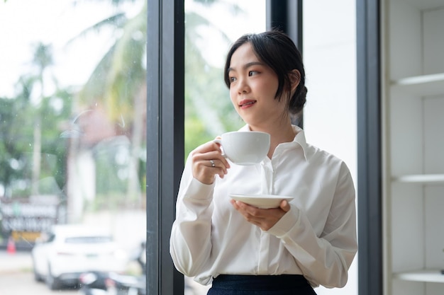 Una hermosa mujer de negocios asiática bebiendo café mientras está de pie junto a la ventana de su oficina