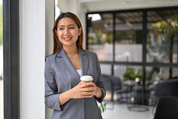 Hermosa mujer de negocios asiática parada junto a la ventana sostiene una taza de café para llevar y sueña despierto