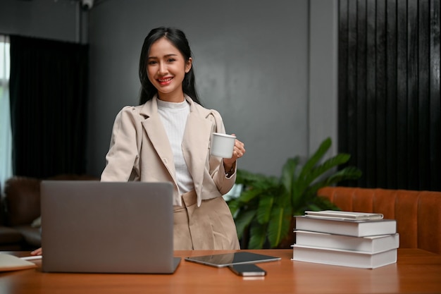 Hermosa mujer de negocios asiática parada en su escritorio sosteniendo una taza de café