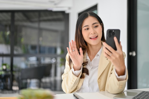 Hermosa mujer de negocios asiática saluda y habla con alguien en una videollamada a través de su teléfono