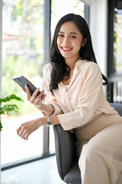 Hermosa mujer de negocios asiática se sienta en un sillón sosteniendo su teléfono inteligente sonriendo y mirando a la cámara