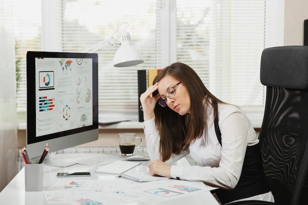 Hermosa mujer de negocios de cabello castaño perplejo y estrés cansada en traje y gafas sentado en el escritorio con una taza de café, trabajando en la computadora contemporánea con documentos en la oficina de luz.