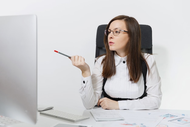 La hermosa mujer de negocios de cabello castaño serio y absorto en traje y gafas sentado en el escritorio, trabajando en la computadora con un monitor moderno con documentos en la oficina de luz,