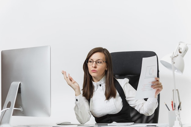 La hermosa mujer de negocios de cabello castaño serio y absorto en traje y gafas sentado en el escritorio, trabajando en la computadora con un monitor moderno con documentos en la oficina de luz,