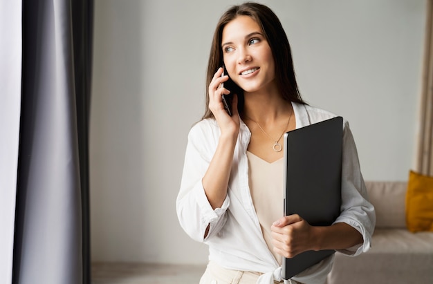 hermosa mujer de negocios con camisa blanca se encuentra en el contexto de la pared blanca junto a la ventana, utiliza el teléfono móvil para el trabajo, sosteniendo la computadora portátil en sus manos.