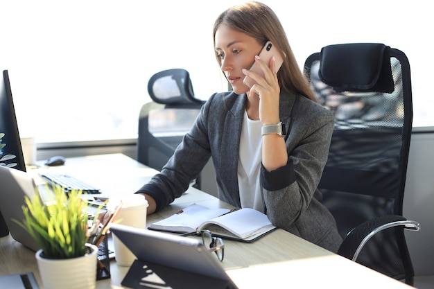 Hermosa mujer de negocios está hablando por teléfono móvil mientras está sentado en la oficina moderna.