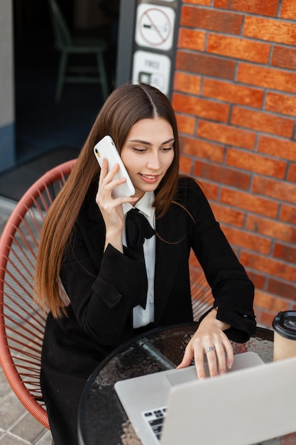 Hermosa mujer de negocios feliz y exitosa en ropa negra de moda con corbata está hablando por teléfono y trabajando en una computadora portátil en la ciudad