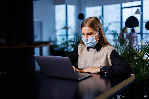 Una hermosa mujer de negocios inteligente en una gran empresa celebra una reunión en línea con socios comerciales con una máscara protectora durante el coronavirus, se sienta en una mesa en un lugar de trabajo con una computadora portátil