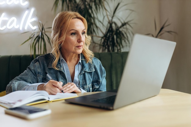 Hermosa mujer de negocios madura que usa una computadora portátil y escribe algo en un cuaderno en el café