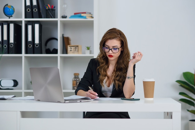 Hermosa mujer de negocios en la oficina con ropa elegante, hace algunas notas en papel mientras está sentado en la oficina moderna y acogedora.