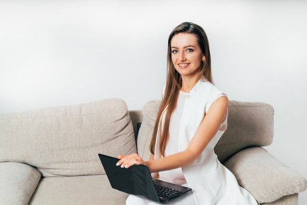 Hermosa mujer de negocios en ropa blanca se sienta en un sofá y sostiene una computadora portátil en su regazo