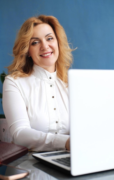 Hermosa mujer de negocios rubia madura sentada en una laptop en la oficina sonriendo y mirando a la cámara