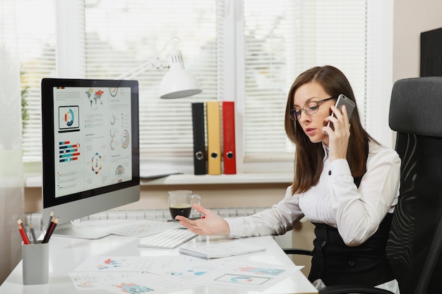 Hermosa mujer de negocios seria en traje y gafas sentado en el escritorio con una taza de café, trabajando en la computadora con documentos en la oficina ligera, hablando por teléfono móvil para resolver problemas, mirando a un lado