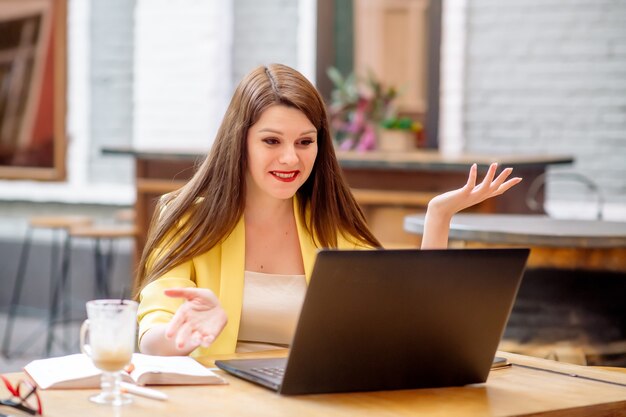 Hermosa mujer de negocios se sienta en una mesa en un café y trabaja de forma remota en una computadora portátil, negociando en línea