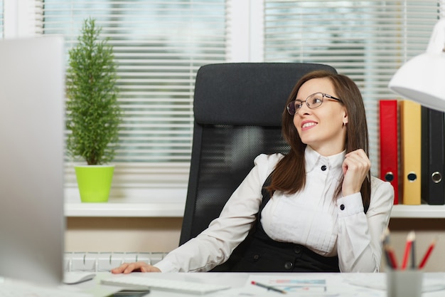 La hermosa mujer de negocios sonriente de cabello castaño con traje y gafas sentada en el escritorio con tableta, trabajando en la computadora con un monitor moderno con documentos en la oficina de luz, mirando a un lado