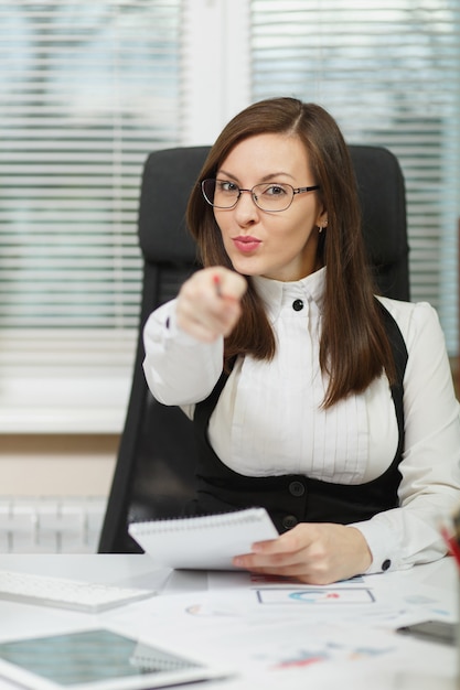 Hermosa mujer de negocios sonriente de cabello castaño con traje y gafas sentado en el escritorio con tableta, trabajando en la computadora con documentos en la oficina de luz, escribiendo con información de lápiz en el cuaderno