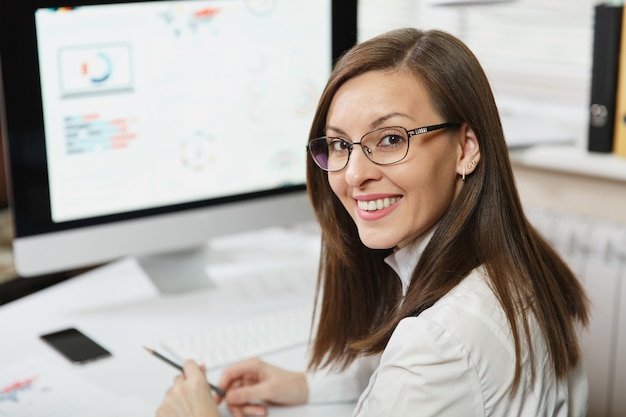 La hermosa mujer de negocios sonriente de cabello castaño en traje y gafas trabajando en la computadora con documentos en la oficina de luz, mirando a la cámara, contra la pared