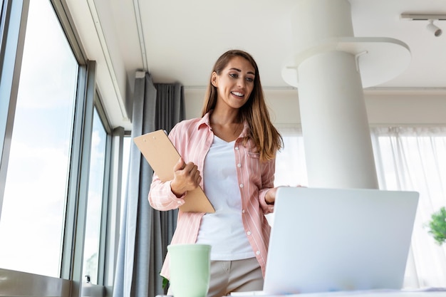 Hermosa mujer de negocios sonriente leyendo algo en una laptop mientras se apoya en la mesa de la oficina